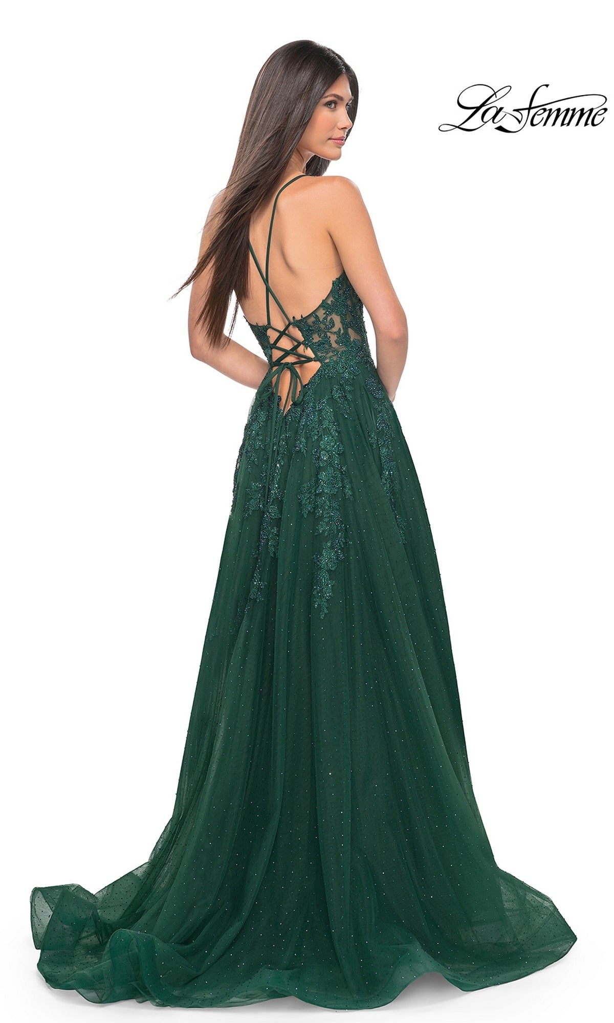 A woman stands looking over her shoulder in the La Femme Lace-Embroidered Long Prom Dress 32147, a dark green gown with floral lace, an open back, and crisscross straps.